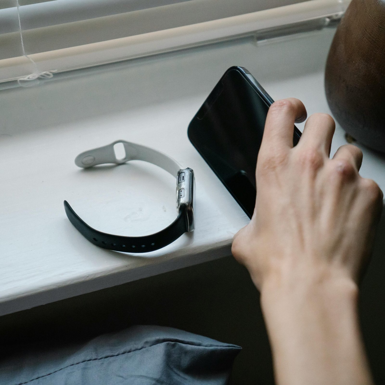 From above of crop anonymous person taking cellphone placed on windowsill near smart watch while waking up in morning